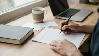 Writer's hand holding a pencil over a manuscript page beside a hardcover book, index cards, coffee and a laptop on a sunlit desk